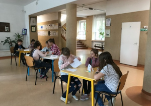 Dzieci rozwiązują przy stolikach w hallu szkoły zadania konkursowe.