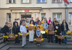 Zdjęcie przedstawia grupę dzieci oraz dwie dorosłe kobiety stojące na schodach przed wejściem do budynku szkoły. Uczestnicy trzymają w dłoniach wiklinowe kosze wypełnione żółtymi żonkilami.
